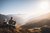 Zwei Personen sitzen auf einem Berg und blicken auf ein sonniges Tal mit Bergen