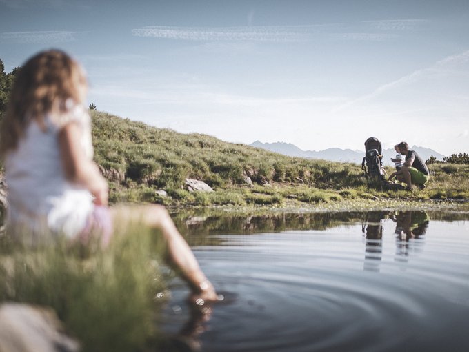 Aktivitäten mit Kindern im Ridnauntal Eine Person sitzt an einem Bergsee, zwei Wanderer rasten am Ufer