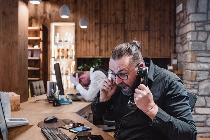 Arbeiten im Erlebnisort Gassenhof Frustrierter Mann telefoniert am Schreibtisch, anderer Mann hält Kopf auf Tisch