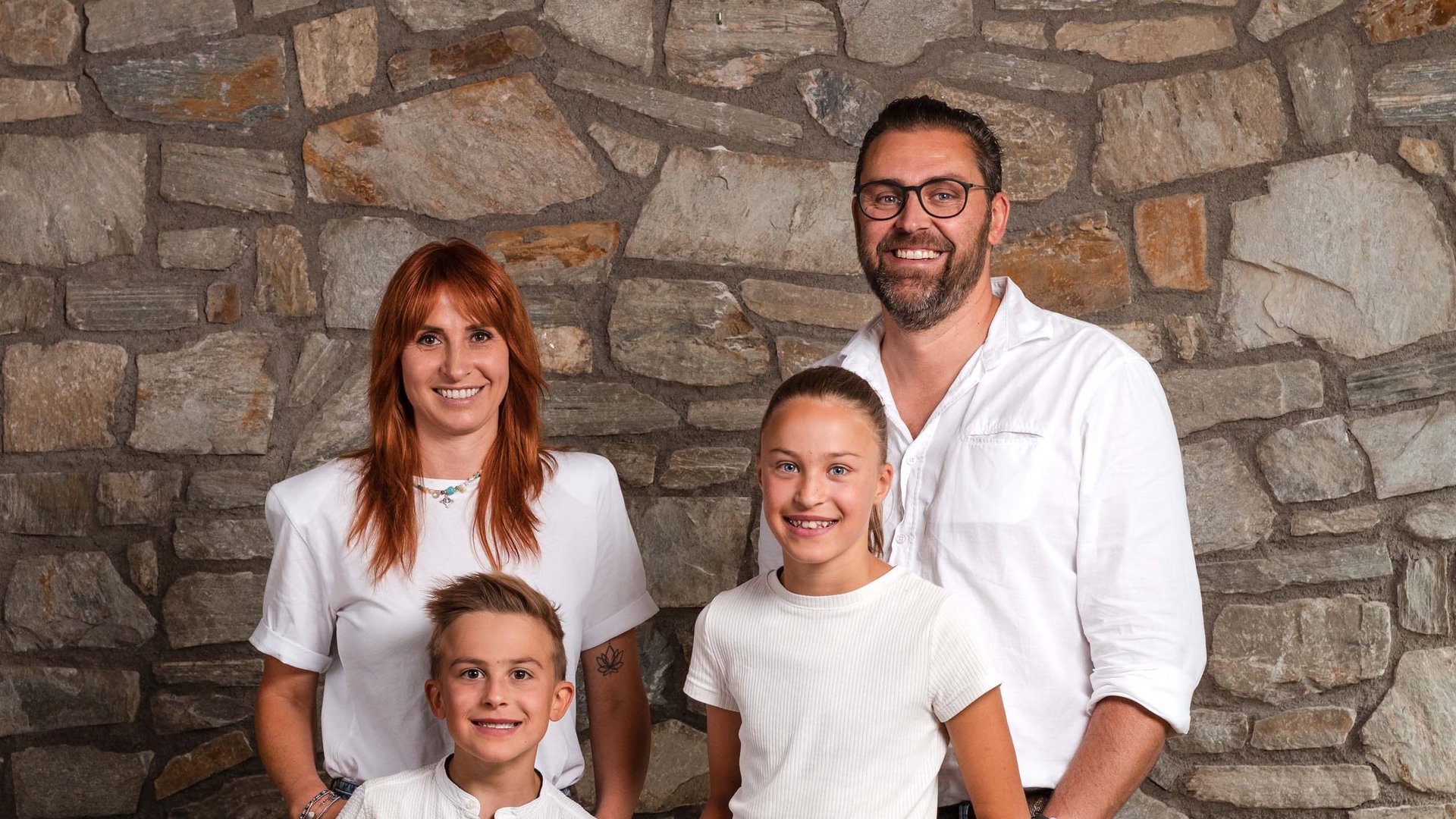 The Volgger family Family of four standing smiling in front of a stone wall, casually dressed