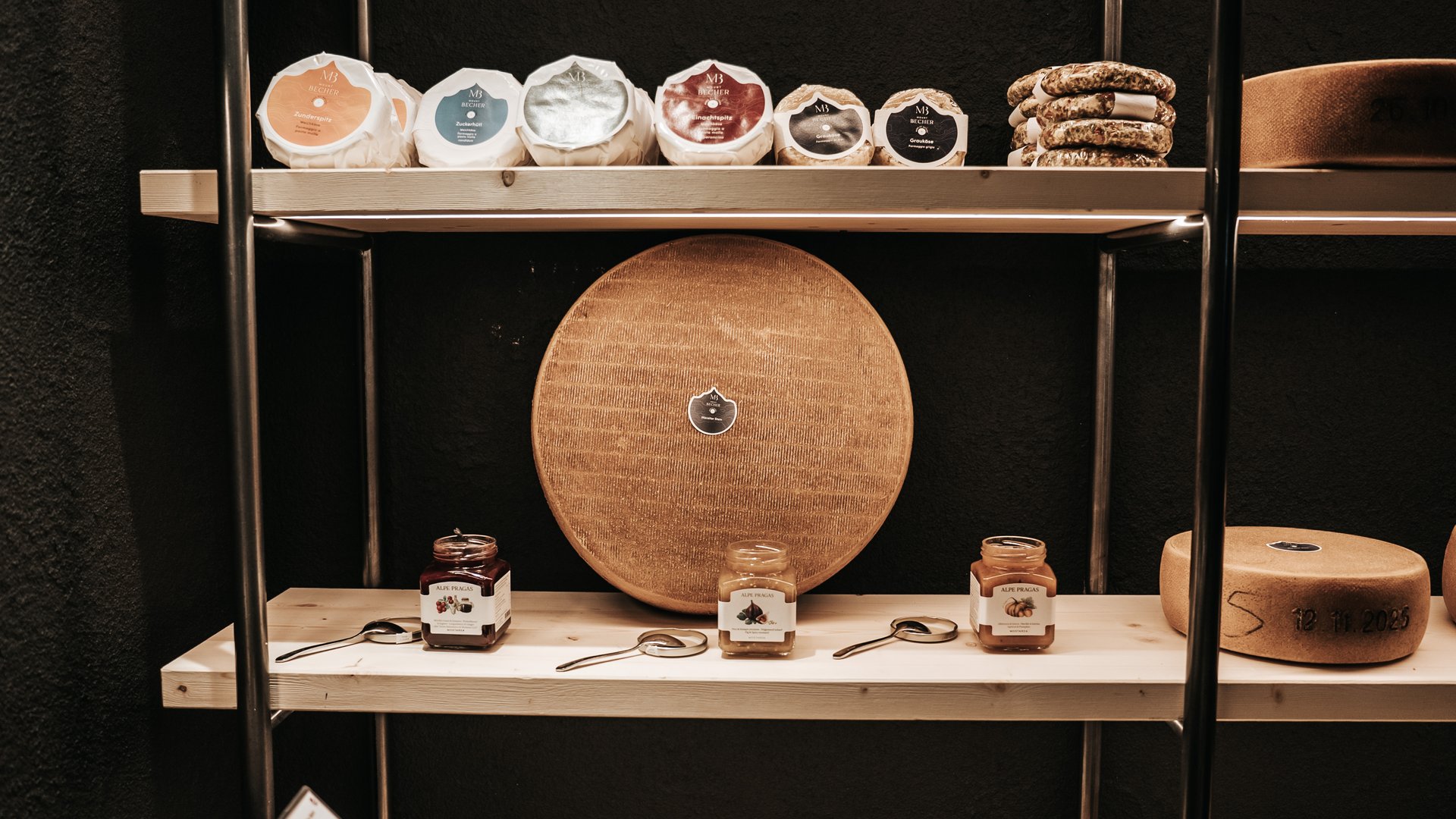 New at Gassenhof Various cheeses and jars of jam on wooden shelves against dark wall