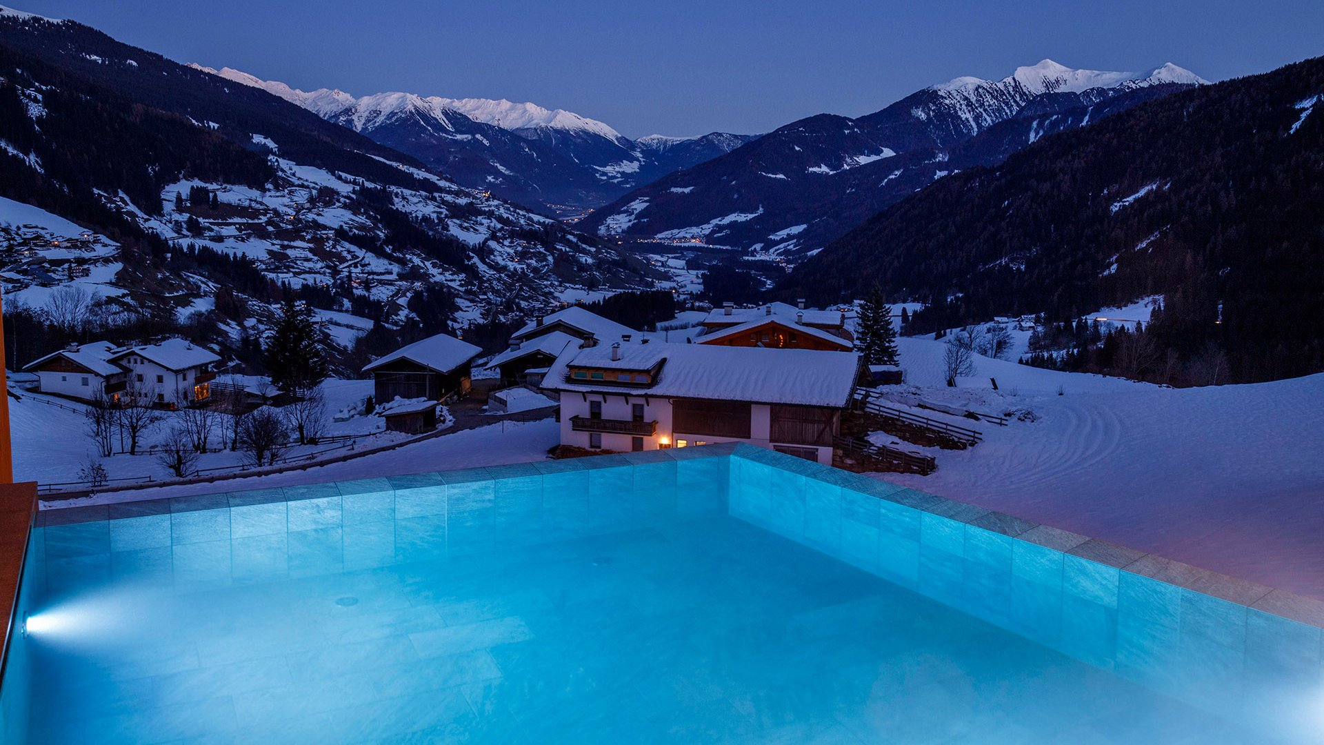 The best spot in the Ridnauntal valley Lit pool overlooking snow-covered Alps and houses at twilight