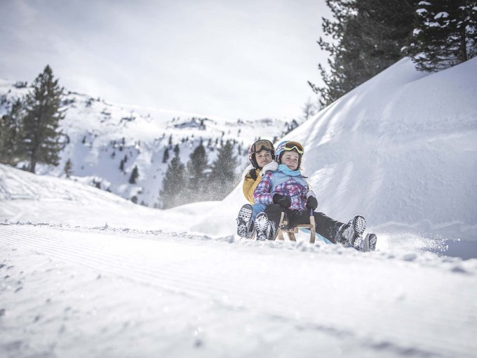 kinder-rodeln-schnee-ratschings Zwei Kinder rodeln zusammen auf einem verschneiten Hang in den Bergen