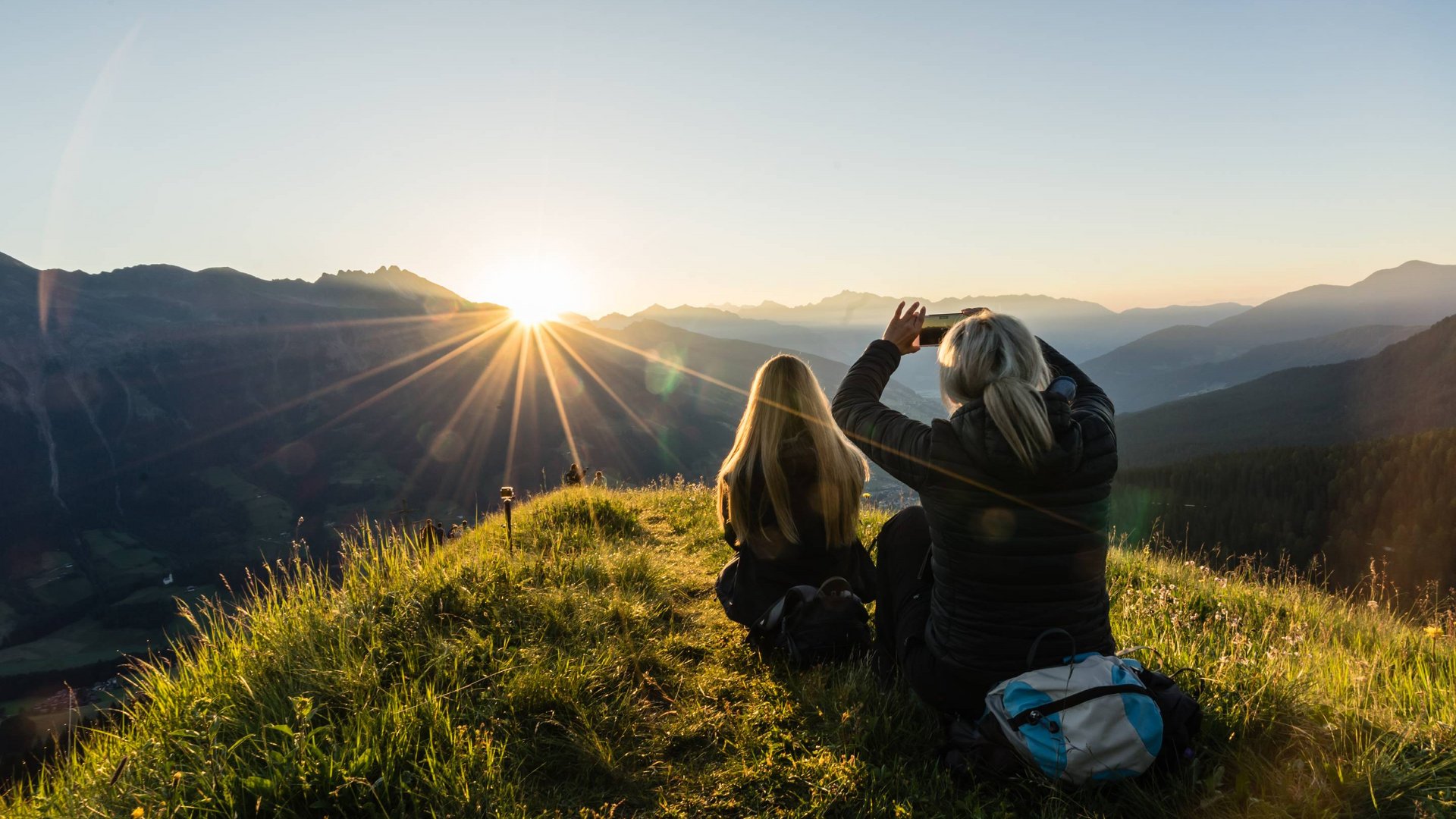 Two people sitting on a hill photographing the sunrise over the mountains