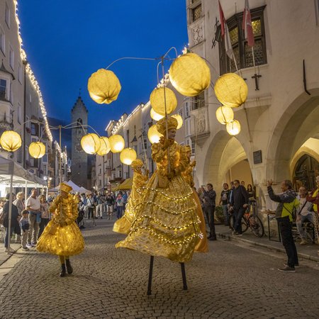 Neuigkeiten Straßenszene bei Nacht mit Menschen in goldenen Kostümen und beleuchteten Kugeln