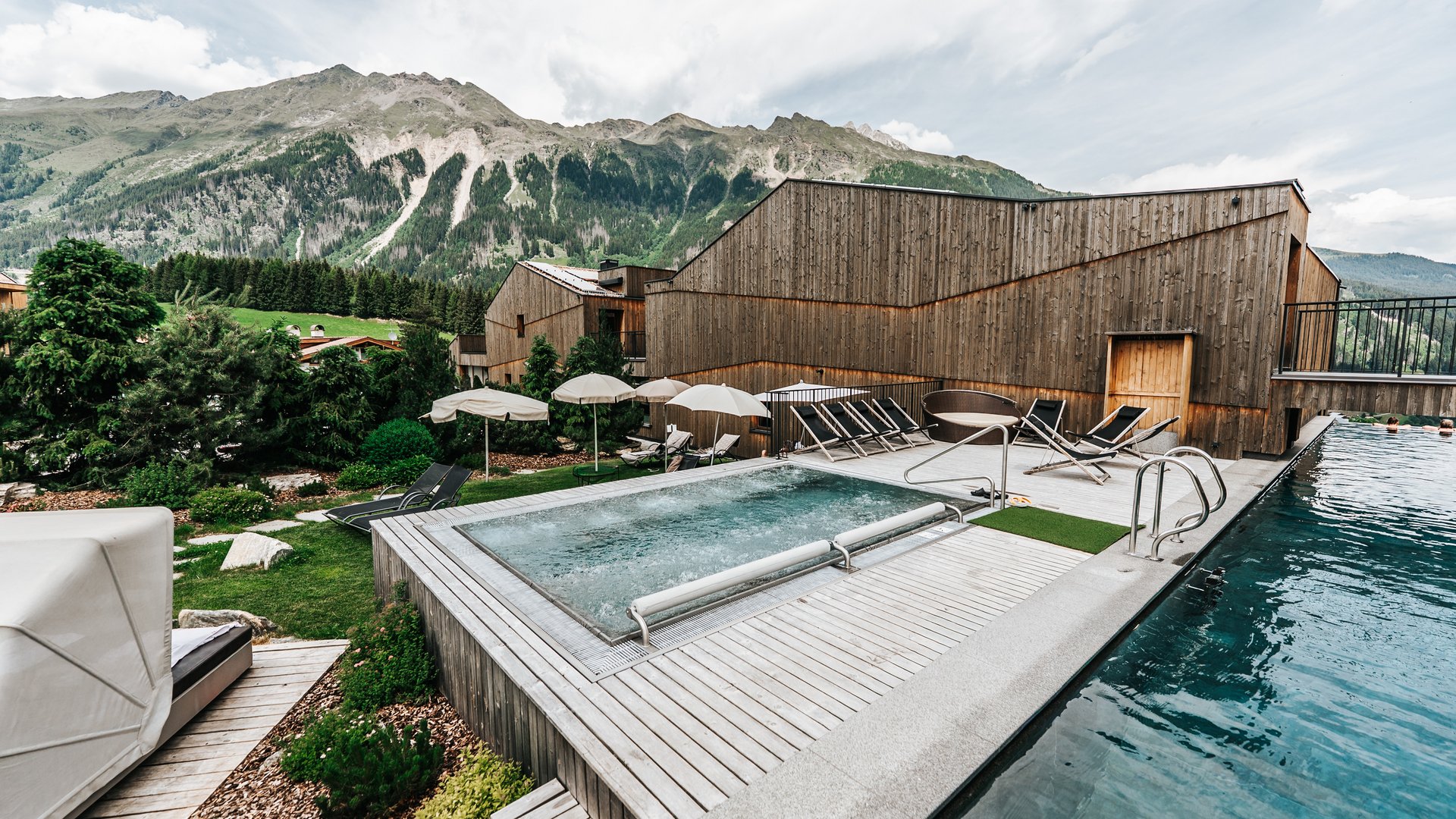 Our water world Outdoor pool and hot tub with lounge chairs by wooden buildings and mountain backdrop