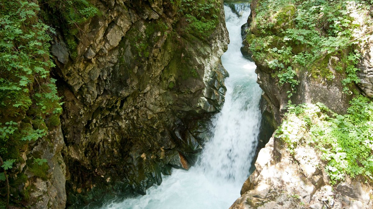 Nachhaltiger Urlaub Wasserfall in einem bewaldeten Tal mit einer Holzbrücke und Spaziergängern