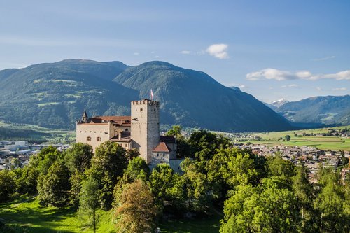 Vacanze in Alto Adige Castello su collina alberata con montagne e città sullo sfondo