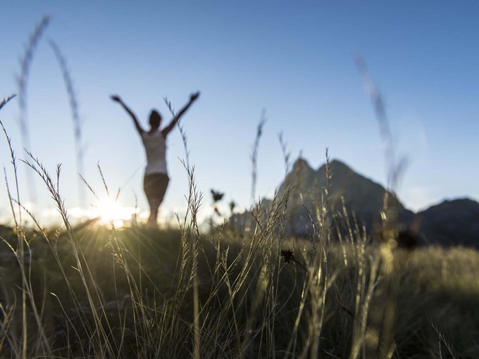 Your base for memorable experiences Person with raised arms in a field at sunset with mountains in the background